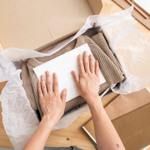 Hands of young female manager of online shop putting paper on top of folded sweater into box before packing it and sending to client